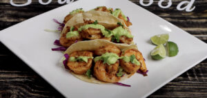 Shrimp tacos topped with creamy avocado sauce and cilantro, served with lime wedges on a white plate.
