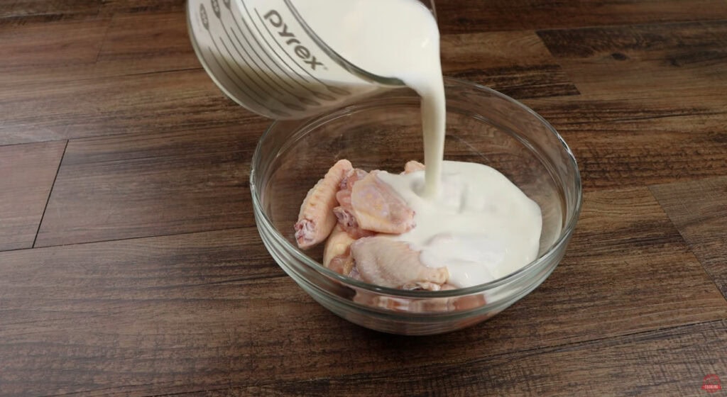 Buttermilk being poured over raw chicken wings in a glass bowl to start the marinade
