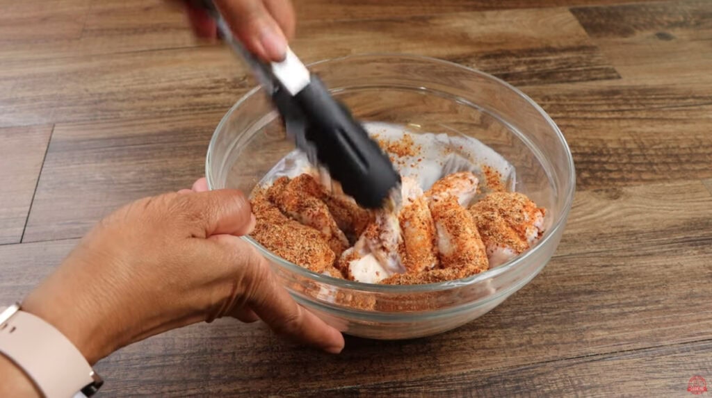  Buttermilk-brined chicken wings being tossed with dry rub seasoning using tongs for Ghost Pepper Wings
