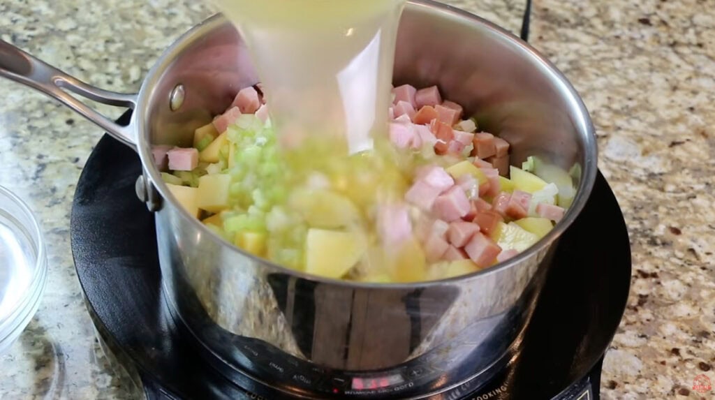 Chicken broth being poured over diced potatoes, ham, celery, and onion in a pot.