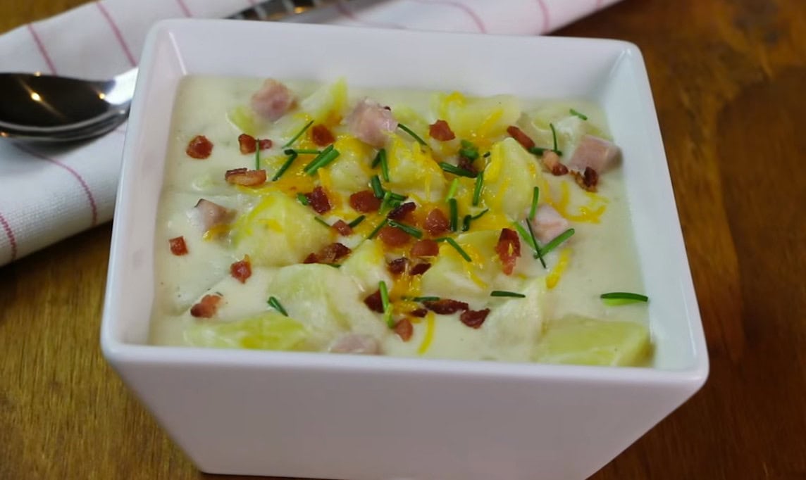 Creamy ham and potato soup in a white bowl topped with bacon, shredded cheddar cheese, and chives.