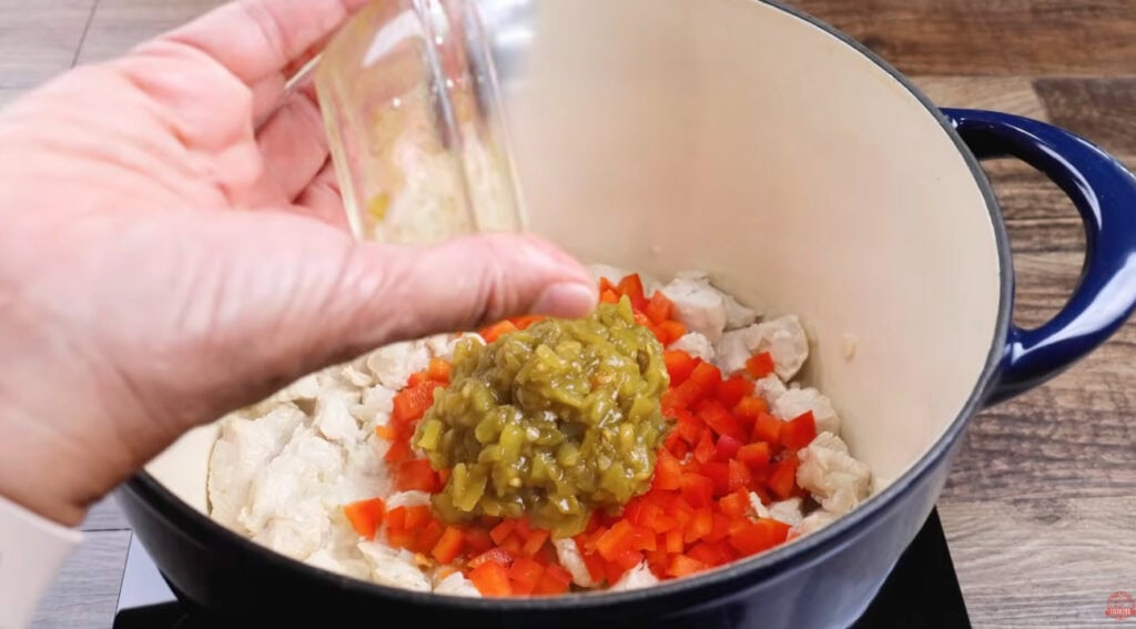 Green chiles being added to chicken and diced red bell pepper in a Dutch oven.