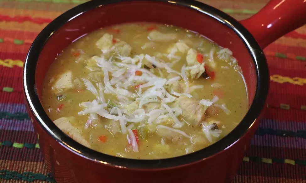 White chicken chili with white beans and red bell pepper served in a red bowl topped with shredded cheese.